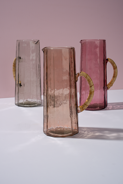 
                    
                      Handblown Hammered Glass Pitchers, light grey, dusky rose, and vibrant pink, with rattan handles, grouped side by side.
                    
                  
