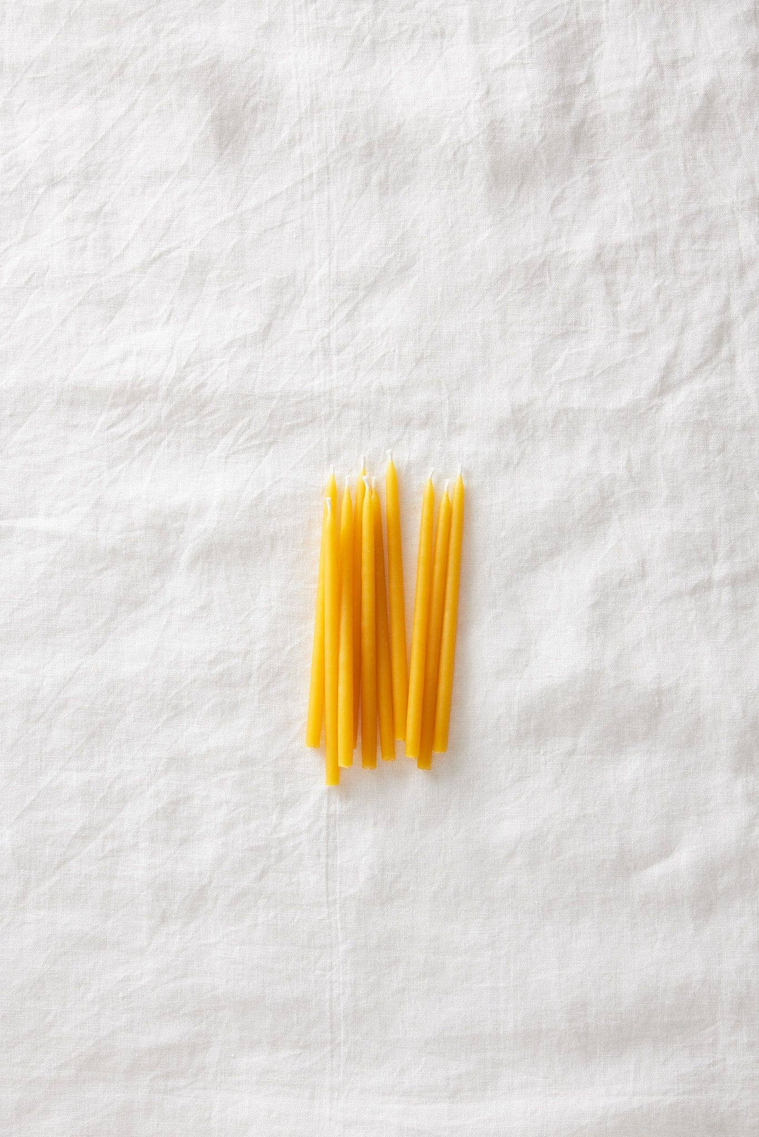 Beeswax Birthday Candles, a loose cluster of yellow pure beeswax candles arranged on a white linen flat lay, top-down view.
