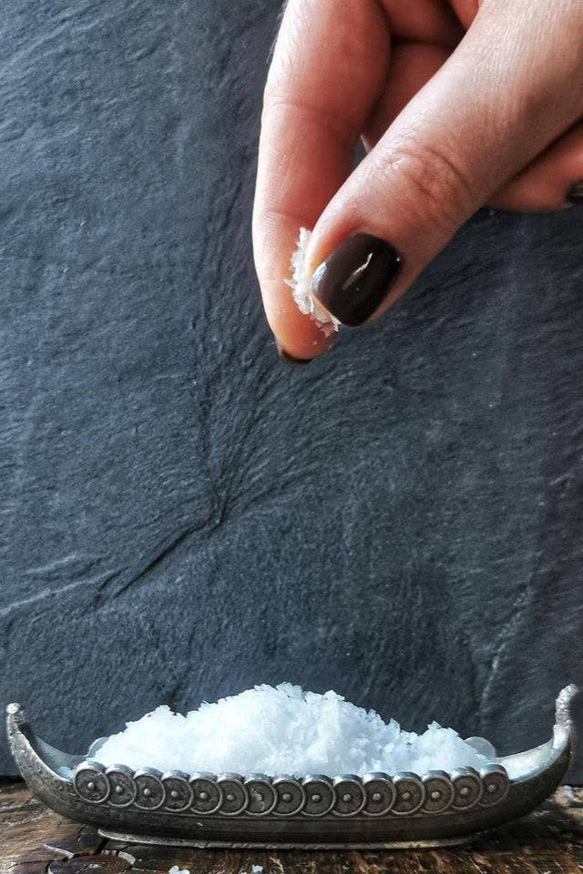 Hand sprinkling Norwegian Sea Salt Flakes from fingertips into a decorative silver dish. Dark nail polish, dark slate…