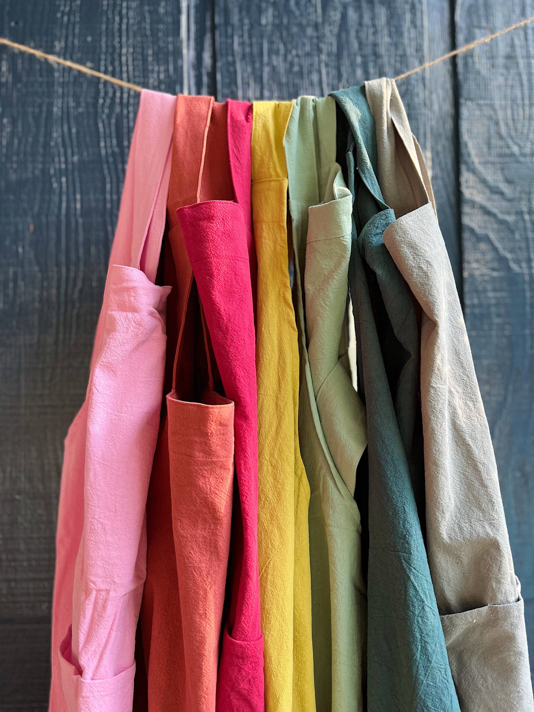 100% Cotton Cross Back Aprons, multi-color array, hanging on rope, showing crinkled fabric texture and pockets.