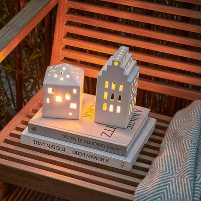 
                    
                      Urbania Town House ceramic candle holder and square companion, glowing warmly, side view on stacked books outdoors.
                    
                  