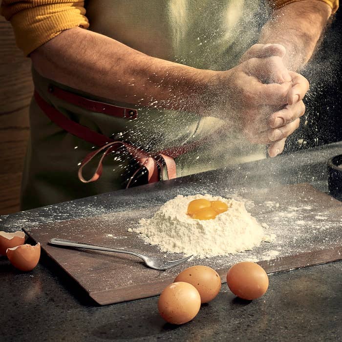 Rå ash wood cutting board, dark, covered in flour and egg yolks for dough preparation, hands dusting flour.