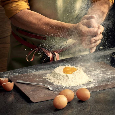 
                    
                      Rå ash wood cutting board, dark, covered in flour and egg yolks for dough preparation, hands dusting flour.
                    
                  