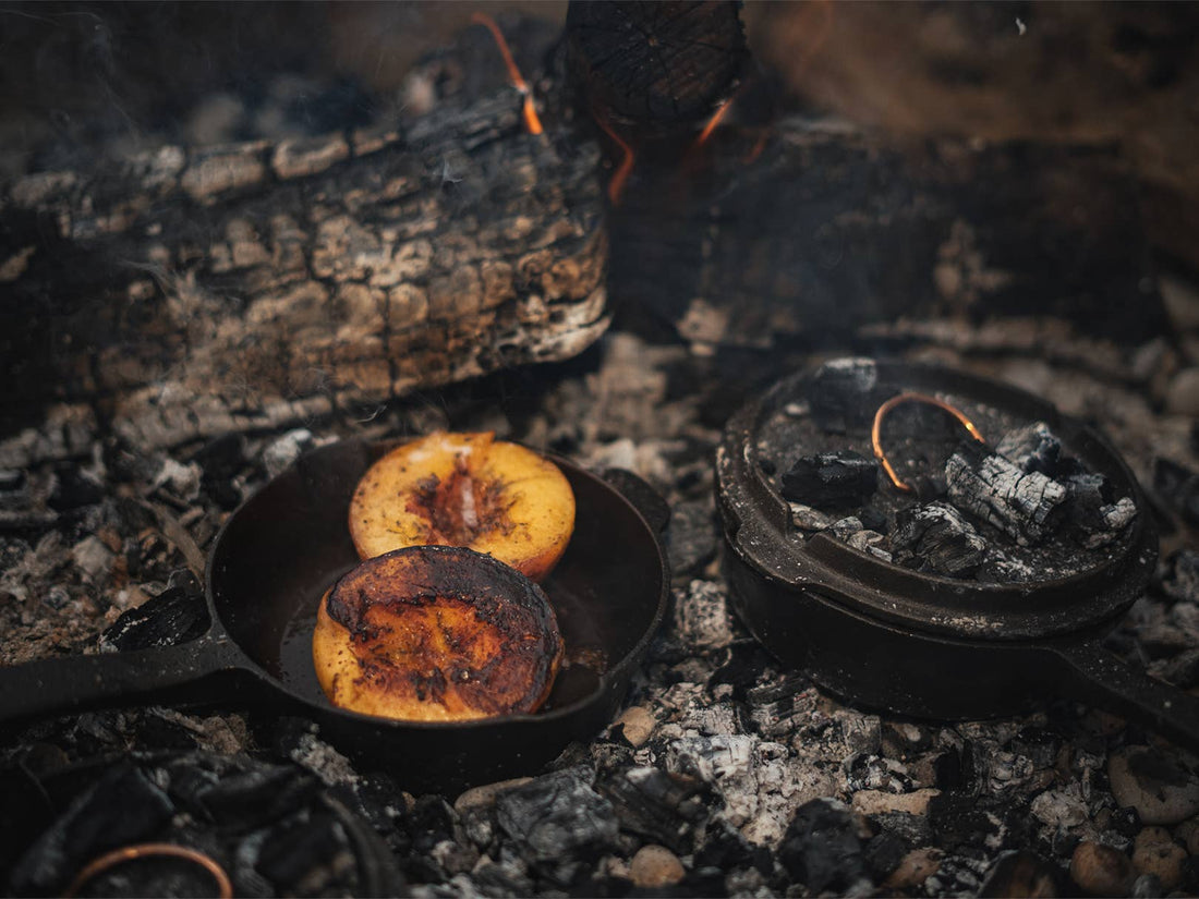 12-inch Cast Iron Skillet cooking peaches over embers, with Dutch oven lid revealing glowing copper handle detail.