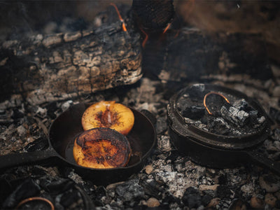 
                    
                      12-inch Cast Iron Skillet cooking peaches over embers, with Dutch oven lid revealing glowing copper handle detail.
                    
                  