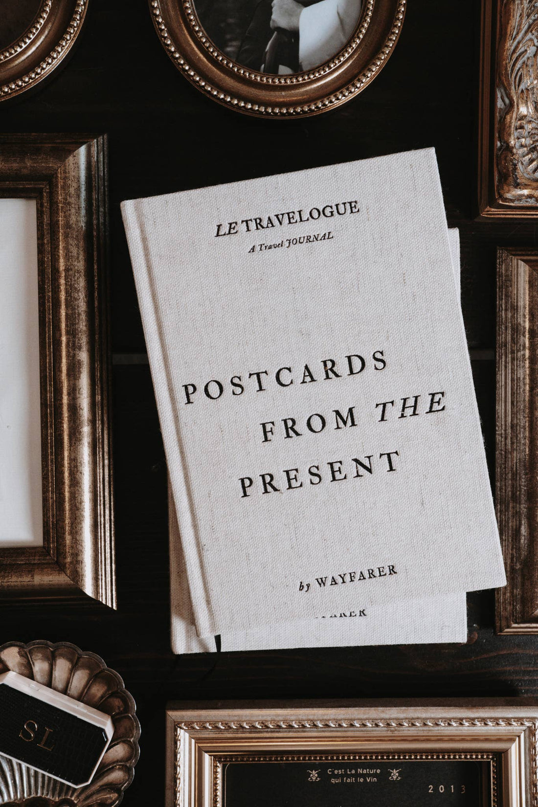 Wayfarer Postcards from the Present travel journal, textured cream linen cover, angled top-down view among gold frames.