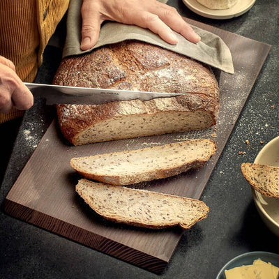 
                    
                      Rå Ash Wood Chopping Board, heat-treated dark ash wood, used to slice bread, with two slices resting on the surface.
                    
                  