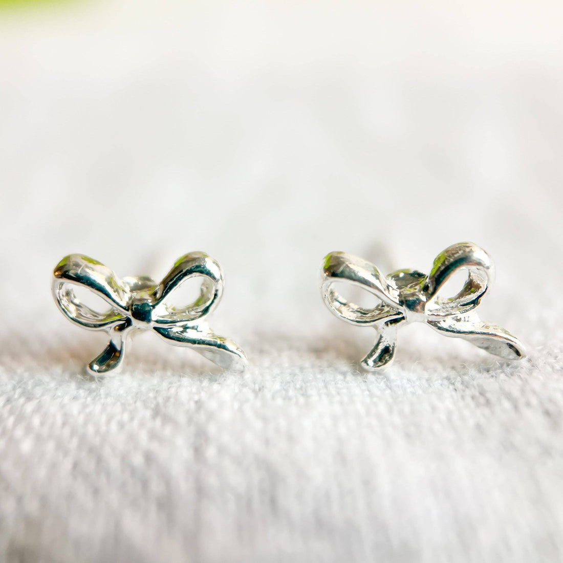 Natalie Clare Collections Mini Silver Bow stud earrings, 925 sterling silver, front view pair on white fabric.