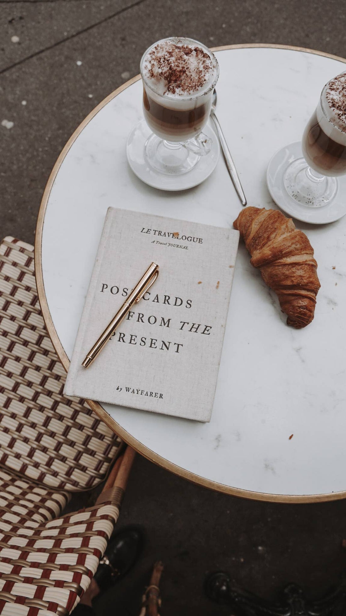 Wayfarer Postcards from the Present travel journal, light linen cover, with gold pen on marble cafe table, croissant and…
