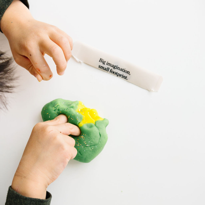 
                    
                      Glitter Natural Play Dough in green and yellow, child's hands mixing with visible gold glitter, top-down view.
                    
                  