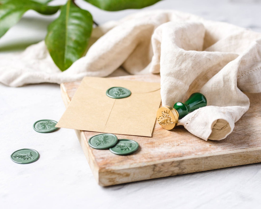 Wax Seal Stamp: gold botanical stamp, green wax seals on kraft envelope, and loose green seals, shown on wood.