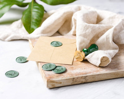 
                    
                      Wax Seal Stamp: gold botanical stamp, green wax seals on kraft envelope, and loose green seals, shown on wood.
                    
                  