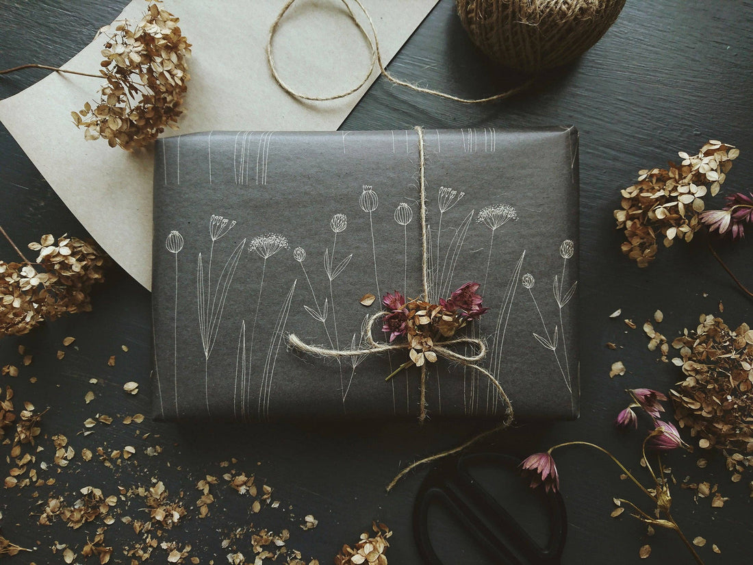 Botanical Wrapping Paper on a gift box, dark gray recycled kraft with white line-art illustrations, tied with twine and…