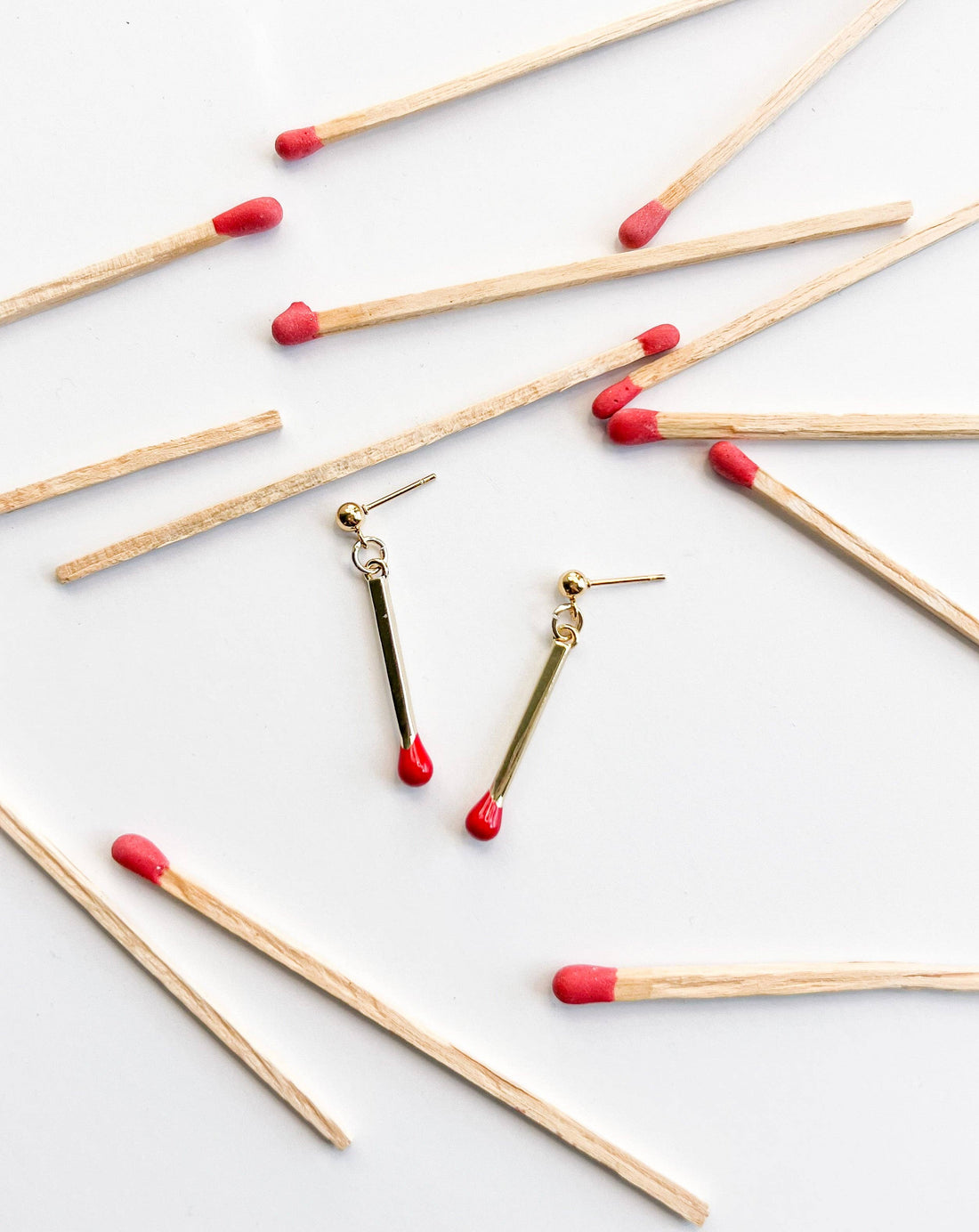 Natalie Clare Collections Matchstick drop earrings, 18k gold plated with red enamel tips, flat lay pair surrounded by wooden…