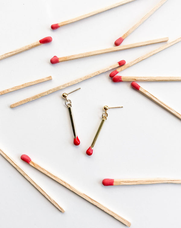 Natalie Clare Collections Matchstick drop earrings, 18k gold plated with red enamel tips, flat lay pair surrounded by wooden…