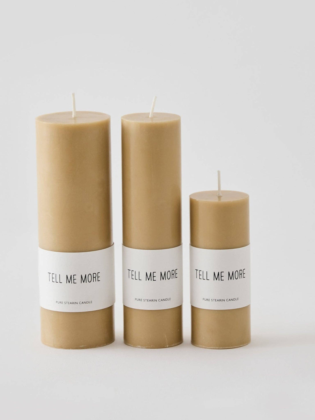 Luna stearin candles, three tan cylinders of different heights with 'Tell Me More' labels, front view.