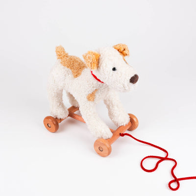 
                    
                      Eliot Dog pull-along toy, white and tan plush, red collar, solid wood wheels, red cotton pull string on floor.
                    
                  