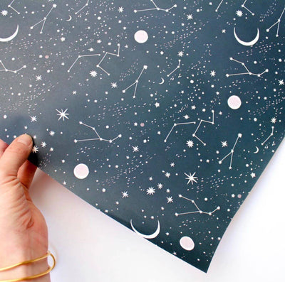 
                    
                      Moon and Stars wrapping paper, dark blue with white constellations, stars, and light pink planets, held in hand for scale.
                    
                  