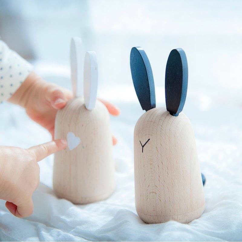 Usagi wooden bunny baby soothers, one with white ears, one with dark blue, child's hands interacting on soft blanket.
