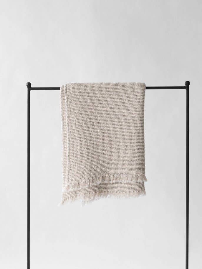 Calma waffle-weave blanket, natural cotton linen blend, draped on a black rail, showing its full texture and fringed ends.