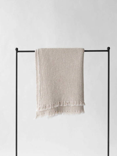 
                    
                      Calma waffle-weave blanket, natural cotton linen blend, draped on a black rail, showing its full texture and fringed ends.
                    
                  