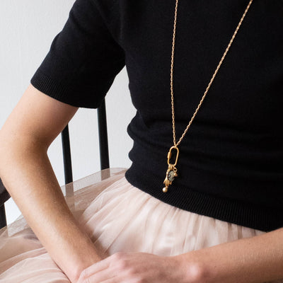 
                    
                      Gold Carabiner Charm Necklace, with cameo, hand, and pearl charms, worn long over a black top.
                    
                  
