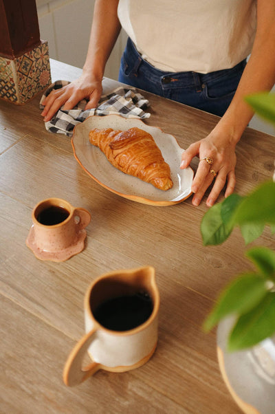 
                    
                      Handcrafted Stoneware Tray, light speckled glaze with natural irregular edge, held in hand with a croissant.
                    
                  