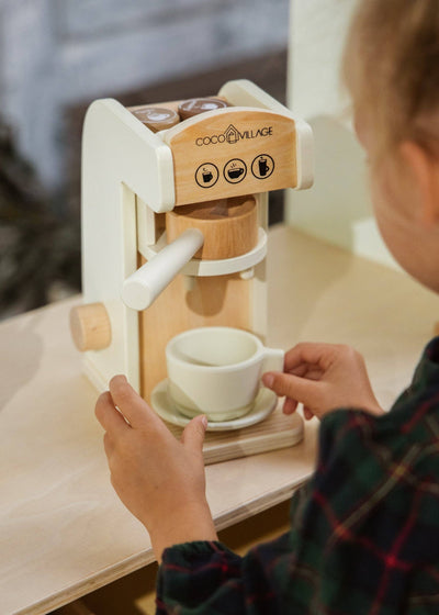 
                    
                      Wooden Coffee Maker Set play kitchen, child's hands holding white cup and saucer, natural wood and white details.
                    
                  