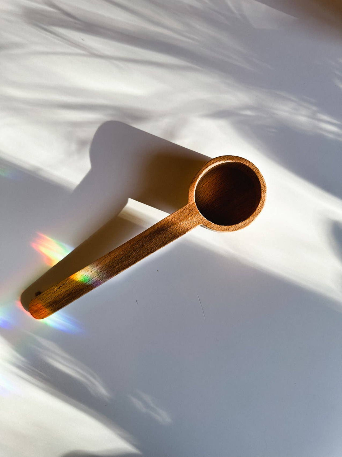 Wooden Tea Scoop with rich dark brown wood grain, diagonal top-down view with dappled light and rainbow prisms.
