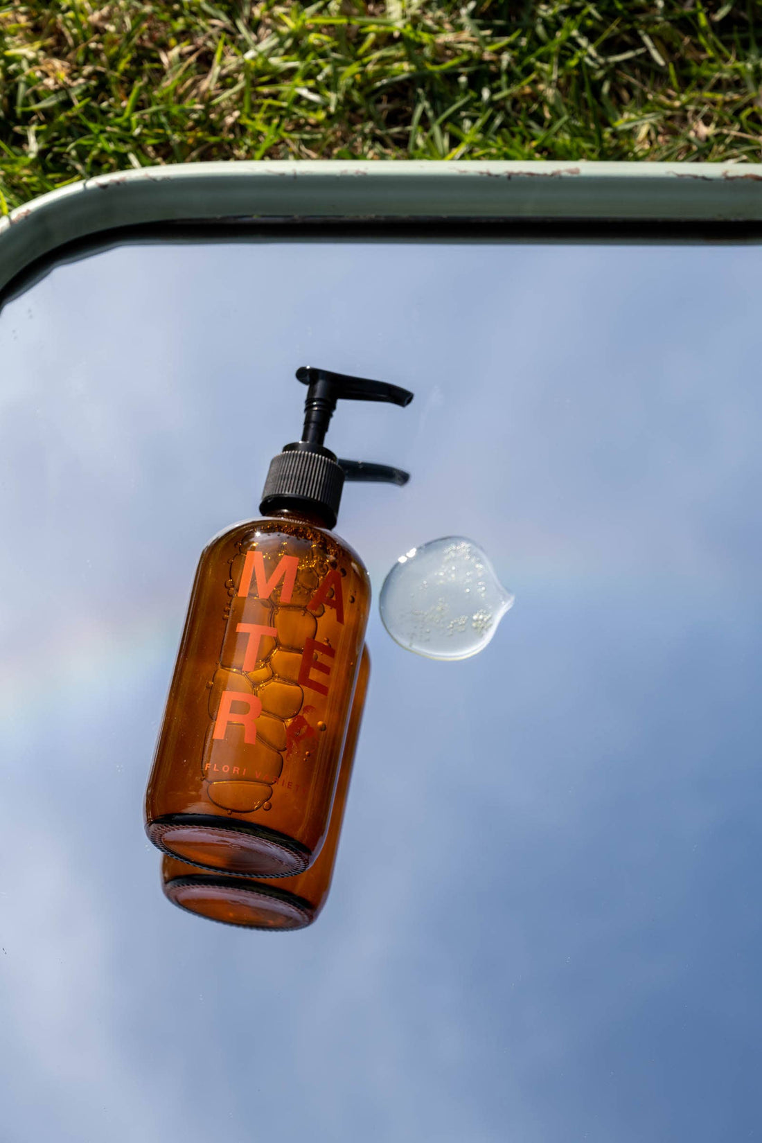 Flori Body and Hand Soap, amber glass bottle with black pump, clear soap droplet visible on mirror, orange logo.