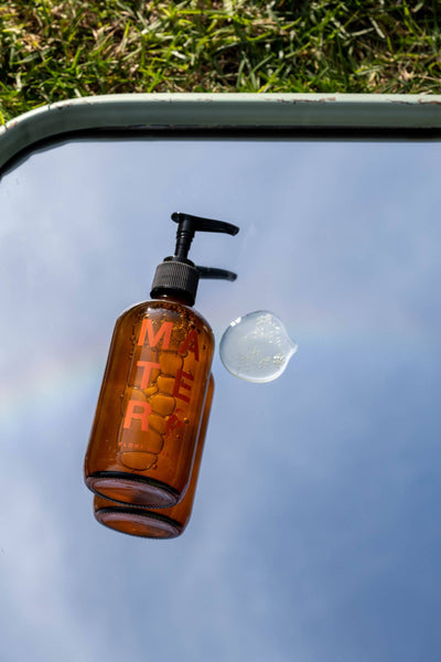 
                    
                      Flori Body and Hand Soap, amber glass bottle with black pump, clear soap droplet visible on mirror, orange logo.
                    
                  