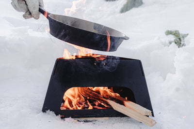 
                    
                      Barebones Flatbrook Folding Stove, black, burning wood in snow, gloved hand holding cast iron pan over flames
                    
                  