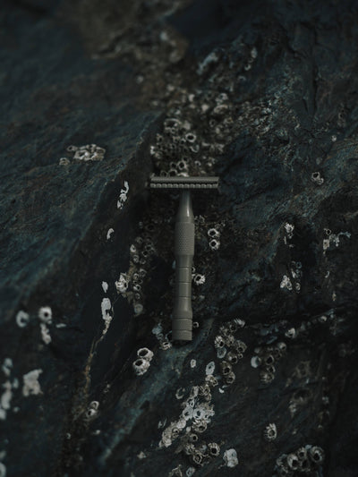 
                    
                      Kelp Brass safety razor, subtle kelp green, close-up view resting vertically on dark rock with barnacles.
                    
                  