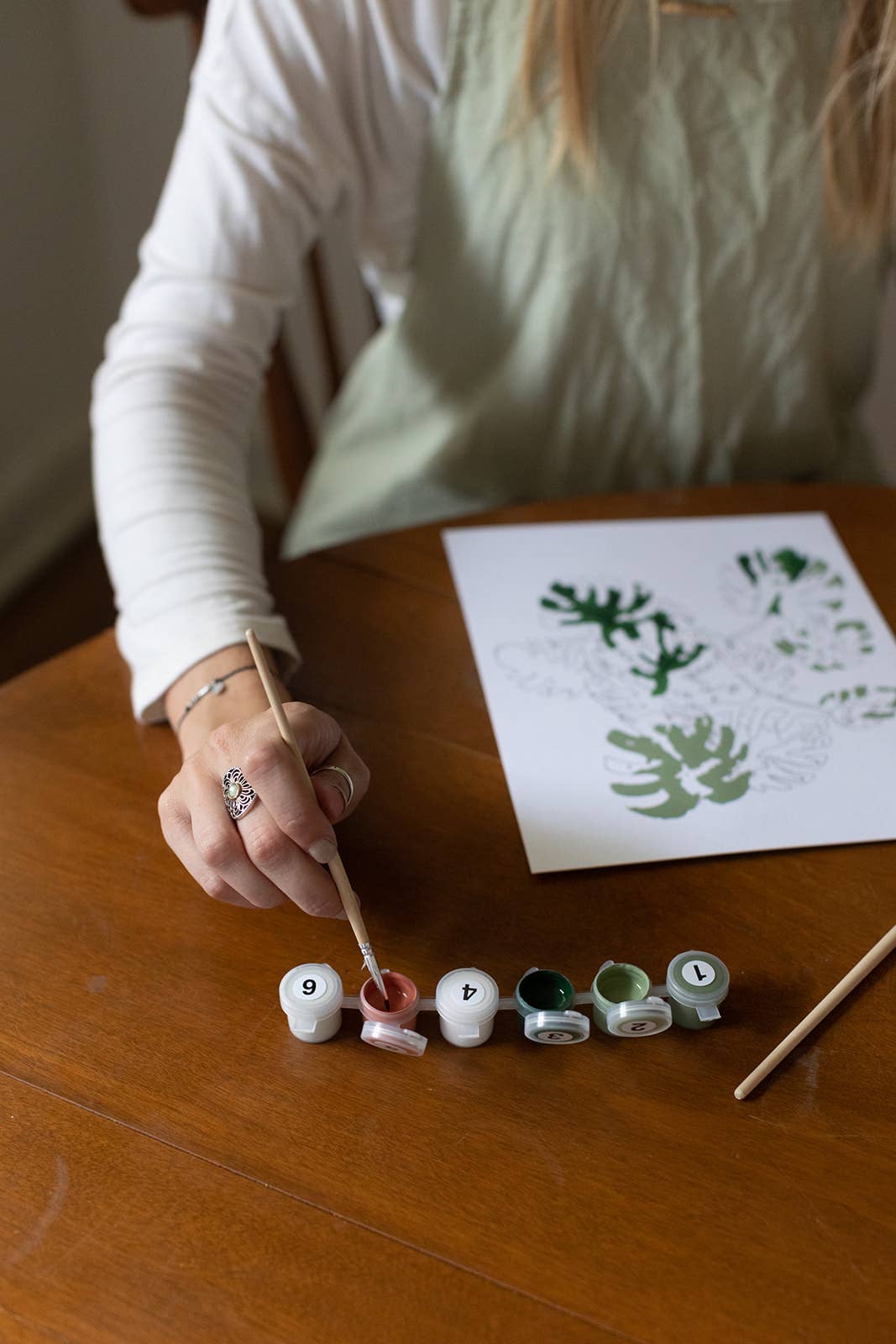 Peony Blossoms Paint-by-Numbers Kit: hand dipping brush into numbered brown paint pot, working on green leaf design.