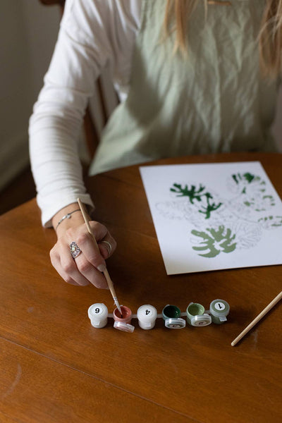 
                    
                      Peony Blossoms Paint-by-Numbers Kit: hand dipping brush into numbered brown paint pot, working on green leaf design.
                    
                  