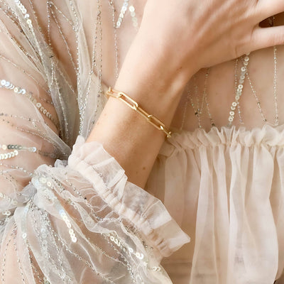 
                    
                      Nest Pretty Things Chunky Gold Paperclip Bracelet, matte gold, worn on wrist over a shimmering sequin sleeve.
                    
                  