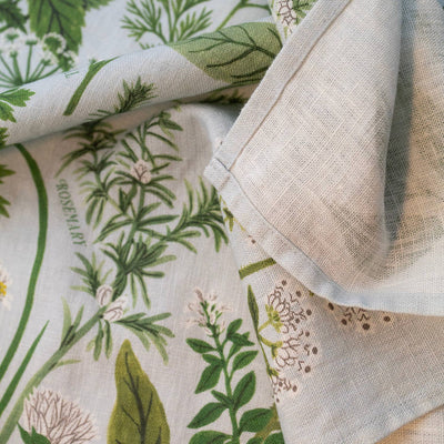 
                    
                      Herb Garden linen tea towel, close-up of folded light blue organic linen fabric, showing hem and botanical herb pattern.
                    
                  
