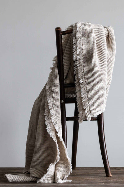 
                    
                      Calma Waffle-Weave blanket in natural cotton-linen, draped over a dark wooden chair, highlighting fringed edges and texture.
                    
                  