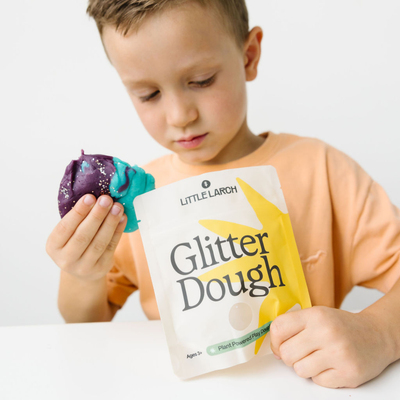 
                    
                      Child holding Little Larch Glitter Dough, purple and teal with glitter, next to its white product package.
                    
                  