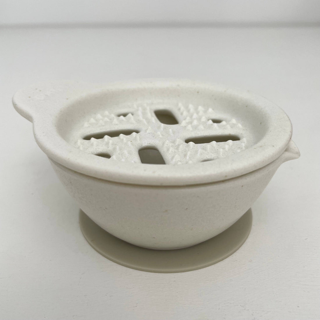 Japanese ceramic vegetable grater, off-white porcelain with textured grating surface, angled side view showing grey silicone…
