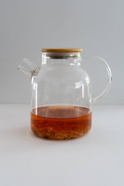 
                    
                      60 Oz. Heat-Resistant Glass Teapot with Bamboo Lid, filled with amber tea and visible loose leaves, side view.
                    
                  
