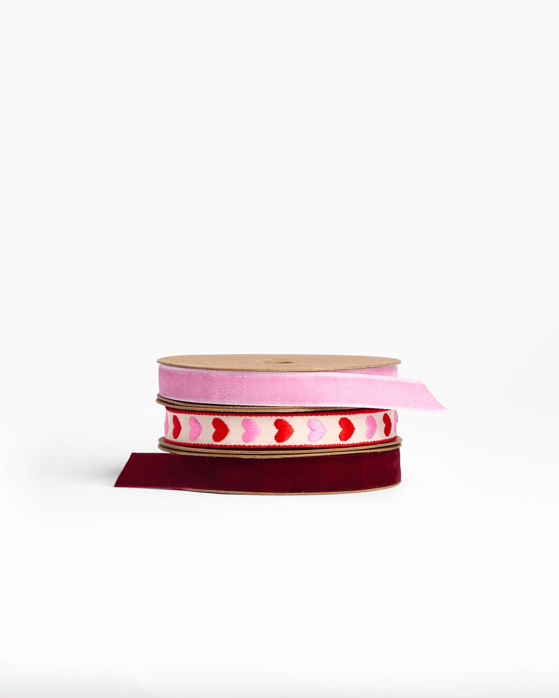 Hearts Woven Ribbon, cream fabric with pink and red embroidered hearts, stacked between pink and burgundy velvet spools.