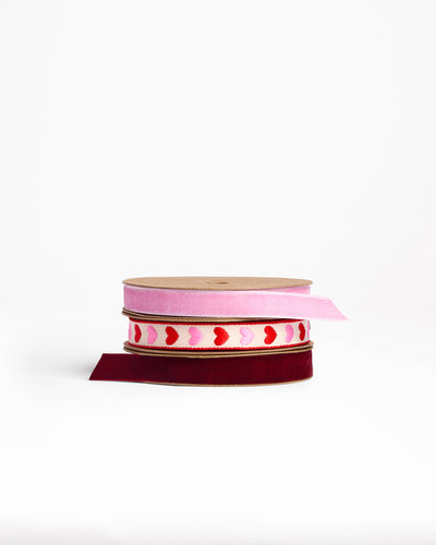 
                    
                      Hearts Woven Ribbon, cream fabric with pink and red embroidered hearts, stacked between pink and burgundy velvet spools.
                    
                  