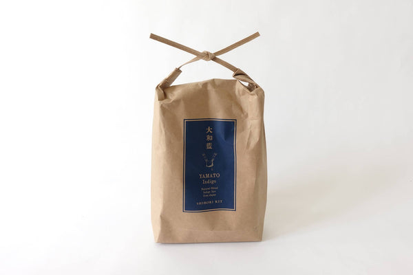 Curious Corners Yamato Indigo Shibori Kit, in a tied kraft paper bag with indigo blue label, full front view on white…