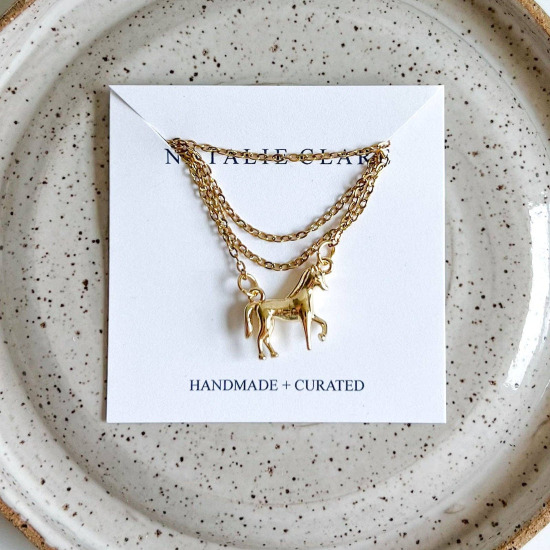 Horse Girl gold necklace with horse pendant, flat lay on its white product card, on a speckled ceramic plate.