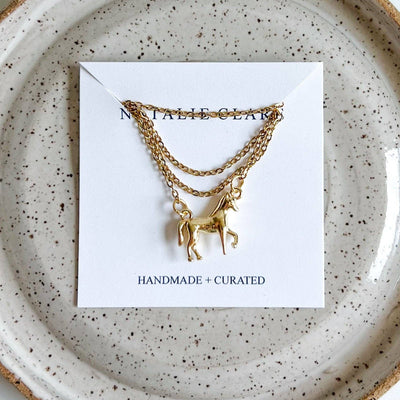 
                    
                      Horse Girl gold necklace with horse pendant, flat lay on its white product card, on a speckled ceramic plate.
                    
                  