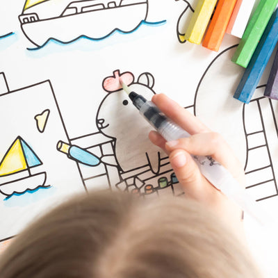 
                    
                      Jumbo Rocket paper coloring book, child's hand colors bear's hat red with a water brush, partially colored boats visible.
                    
                  