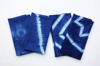 
                    
                      Yamato Indigo Shibori Kit results: four unique cotton fabric pieces with blue and white tie-dye patterns, flat lay.
                    
                  