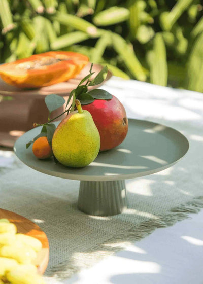 
                    
                      Avisa cake stand, sage green powder-coated metal, close-up showing fluted base detail with fruit.
                    
                  