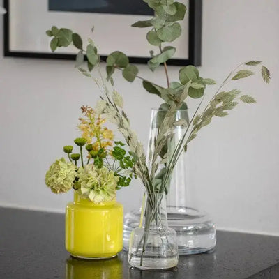 
                    
                      Clear Flora Mouth-Blown Glass Vase holding eucalyptus, in background behind smaller yellow and clear vases on a dark surface.
                    
                  
